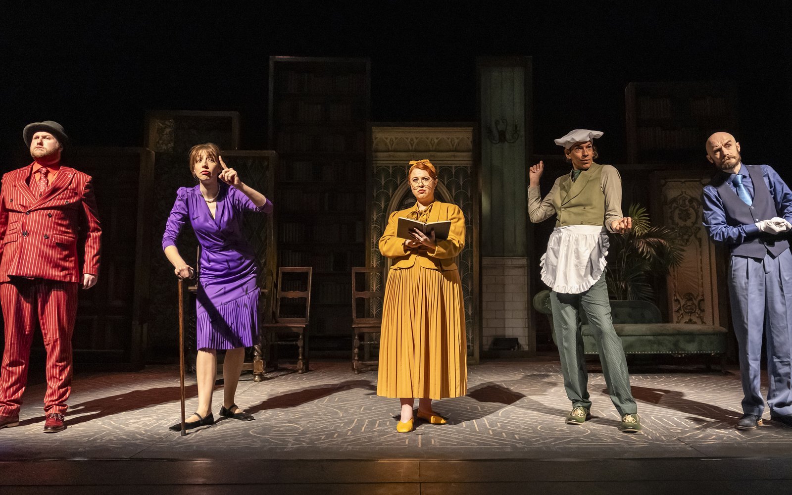 5 people stand in a line, with spotlights on them: a man in a red suit, a woman in a purple dress with a cane, a woman in a yellow skirt with a book, a man dressed in green wearing an apron, and a man in a blue waistcoat.