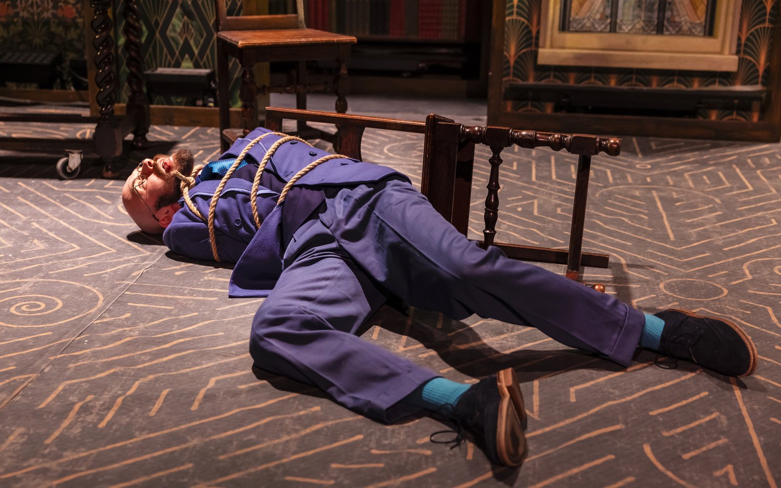 A man in a blue suit lies on the floor. He is tied up with rope. Next to him is a chair that has fallen over.