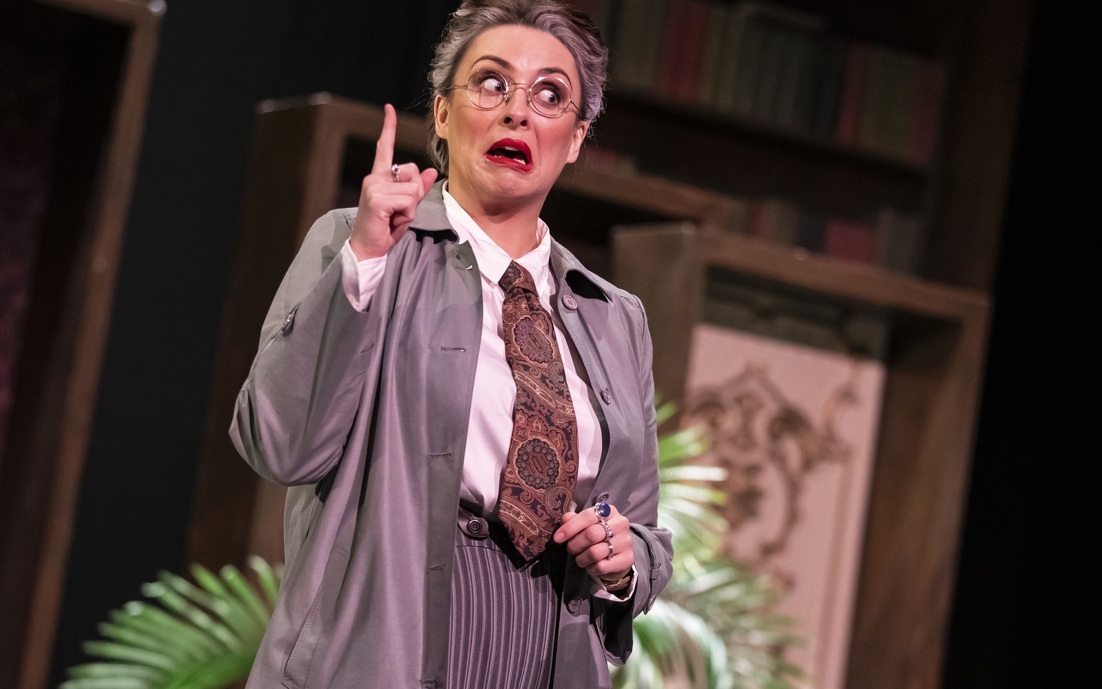 A woman is standing with one finger raised. She is wearing a white shirt, paisley tie, striped trousers, grey overcoat and glasses.