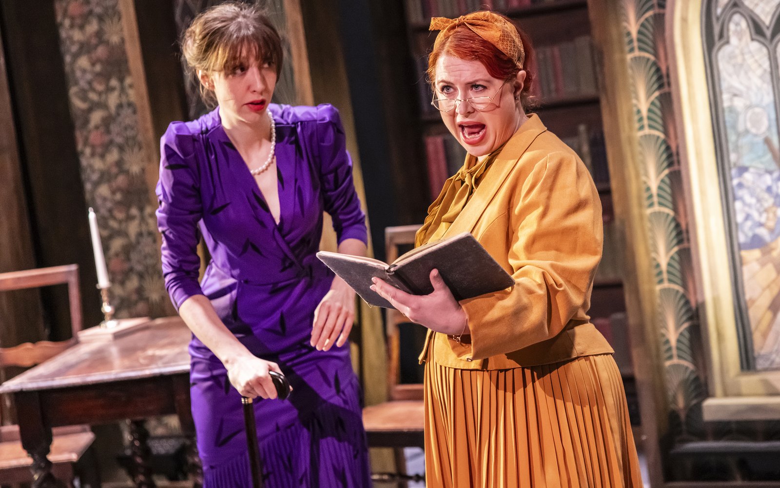 A woman dressed in yellow reads out loud from a book. A woman in a purple dress stands to her left, holding a cane.