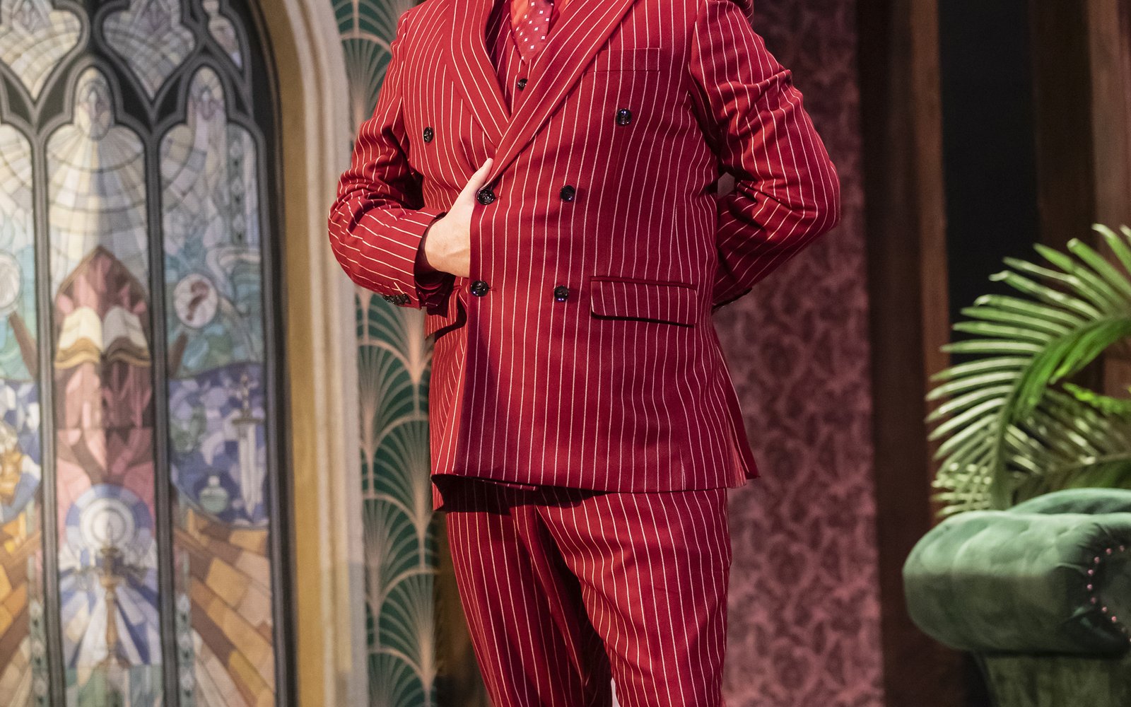 A man stands in a red pinstripe suit. He has one eyebrow raised and one hand tucked into his jacket.