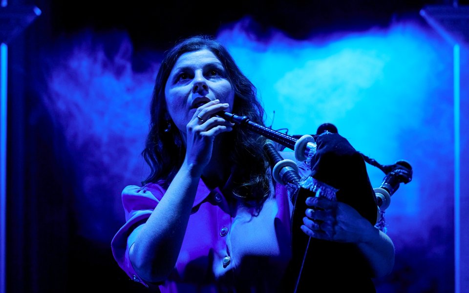 A woman stands in front of a smokey blue background, holding a bagpipe up, preparing to play.