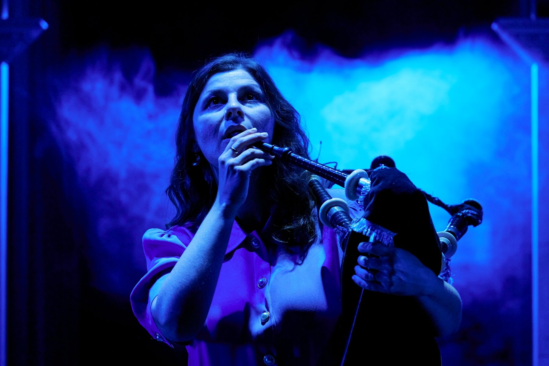 A woman stands in front of a smokey blue background, holding a bagpipe up, preparing to play.