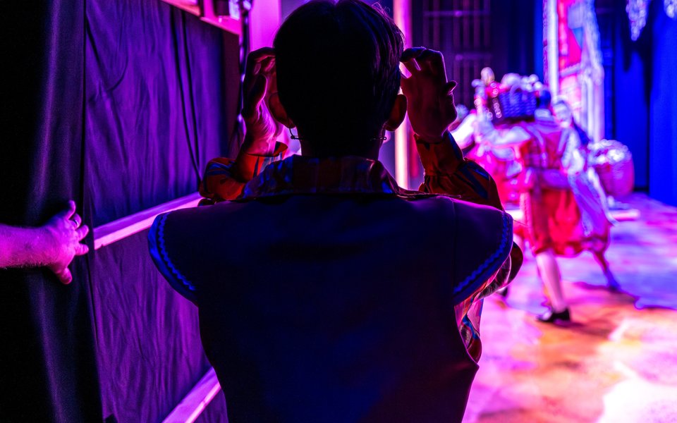 Happy Harry waits in the wings, facing towards the stage. He is fixing his hair. The background is blurry.