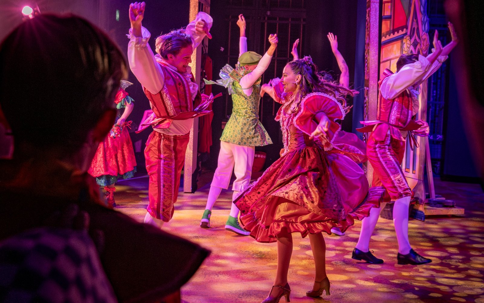 4 performers on stage, seen from the wings. They are dancing, and are all mid-spin. They wear pink medieval costumes.