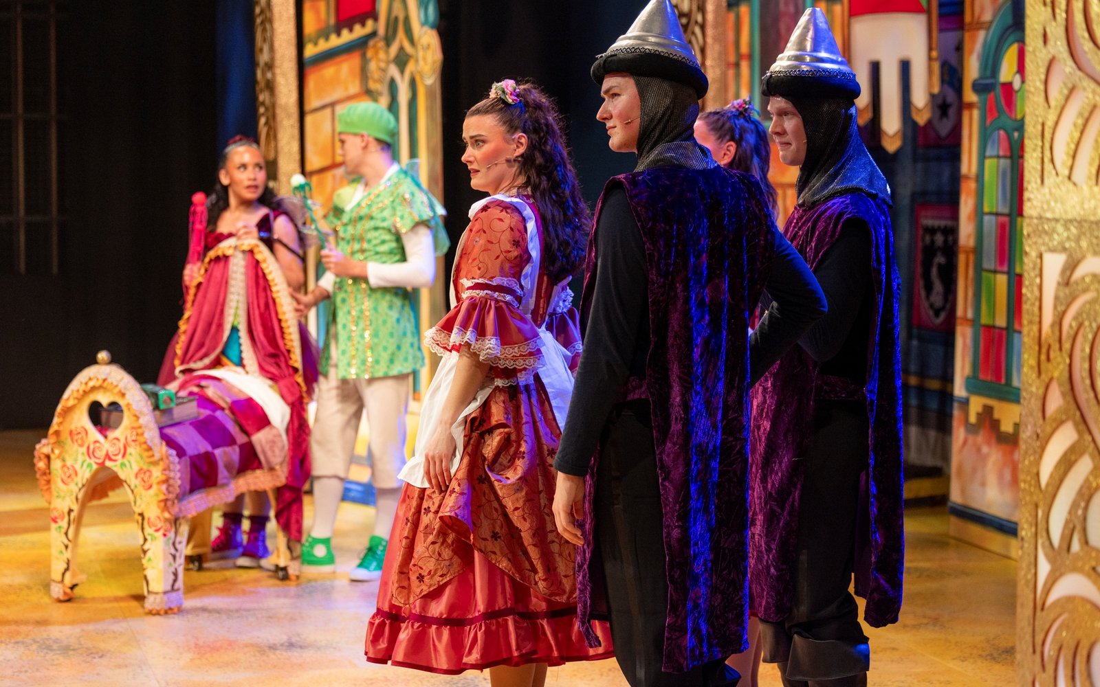 A performer in a pink dress and white apron has a concerned look on her face. Stood next to her are two performers dressed as guards, and in the background are two performers dressed as fairies by an ornate crib.