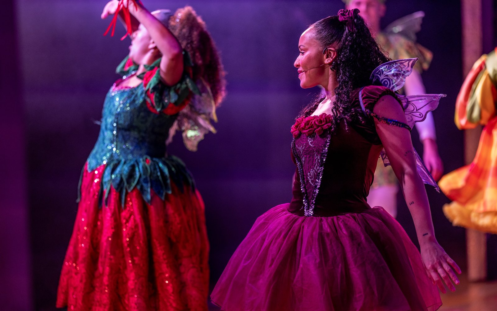 Raspberry the Sprite, wearing a burgundy tutu dress and fairy wings, strikes a pose looking out at the audience. In the background, Fairy Strawberry has her arms raised, like she is in the middle of casting a spell.