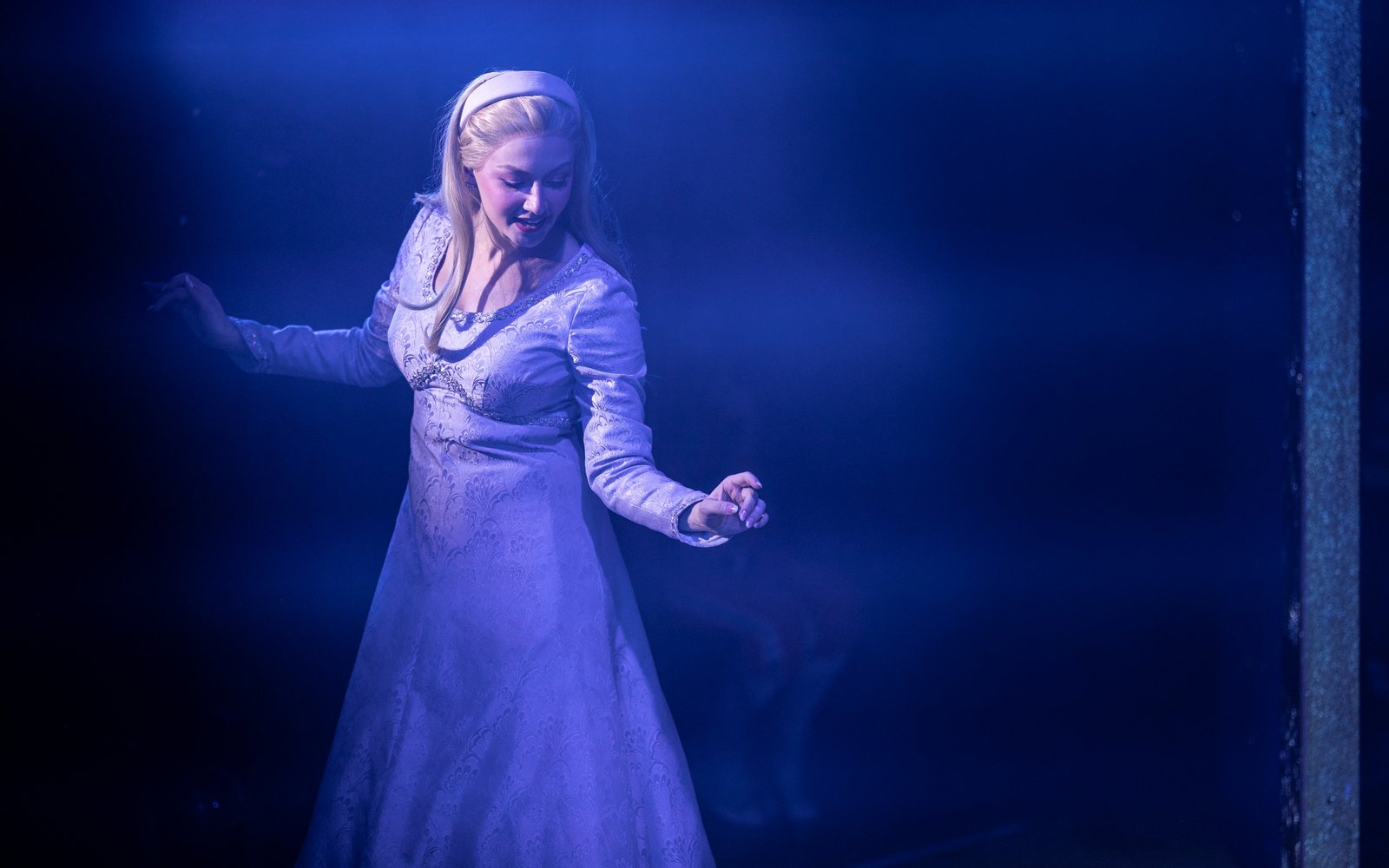 Princess Rose smiles down at the ground. She has blonde hair which is pushed back with a blue hairband, and is wearing a blue jacquard dress.