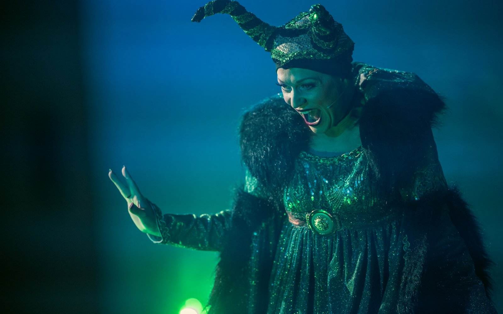 Carabosse, wearing green sparkly horns, a green sparkly dress and a black fur coat, looks delightedly towards the audience.