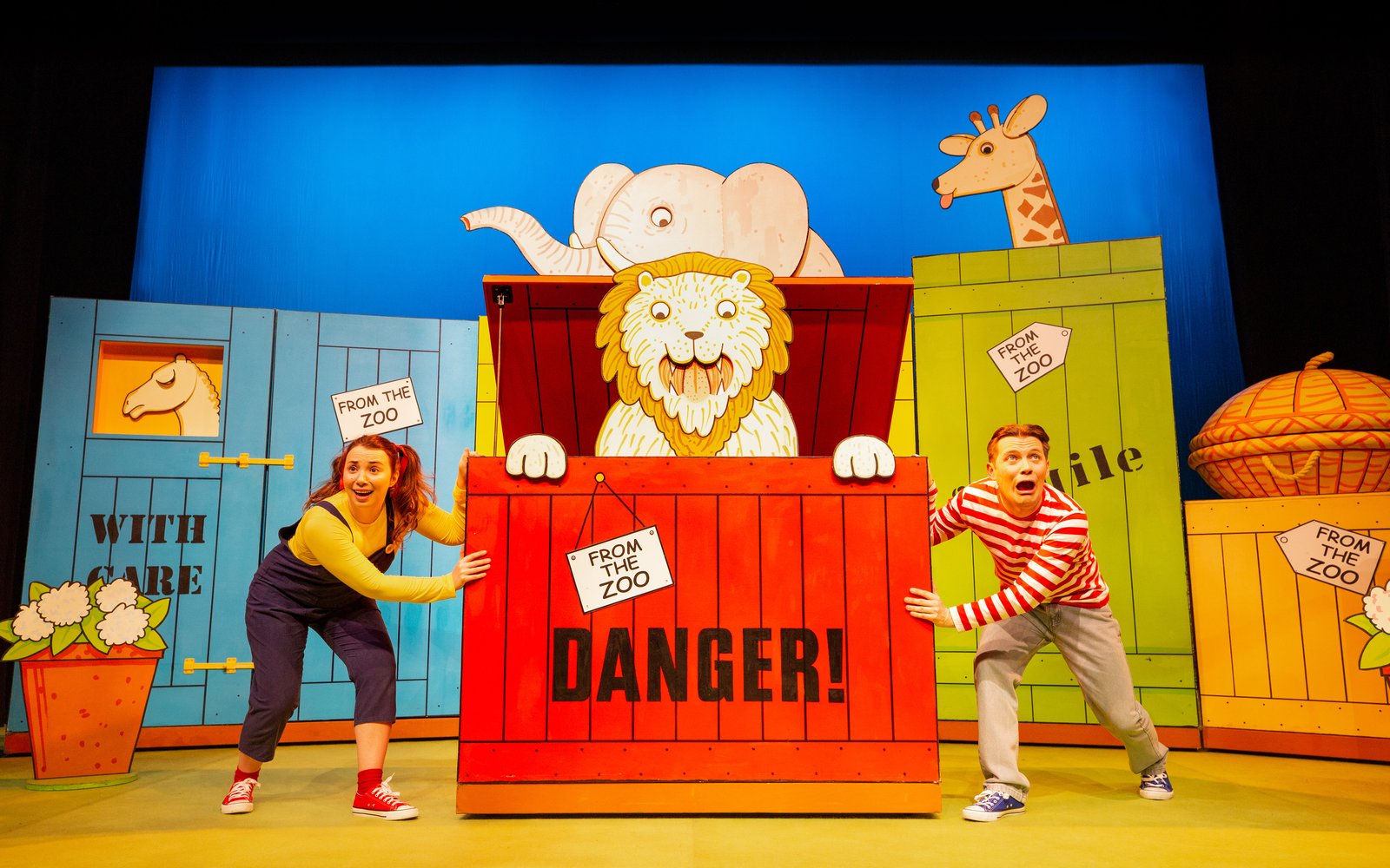 A boy and a girl stand either side of a big red crate, a cartoon lion is sitting inside the box