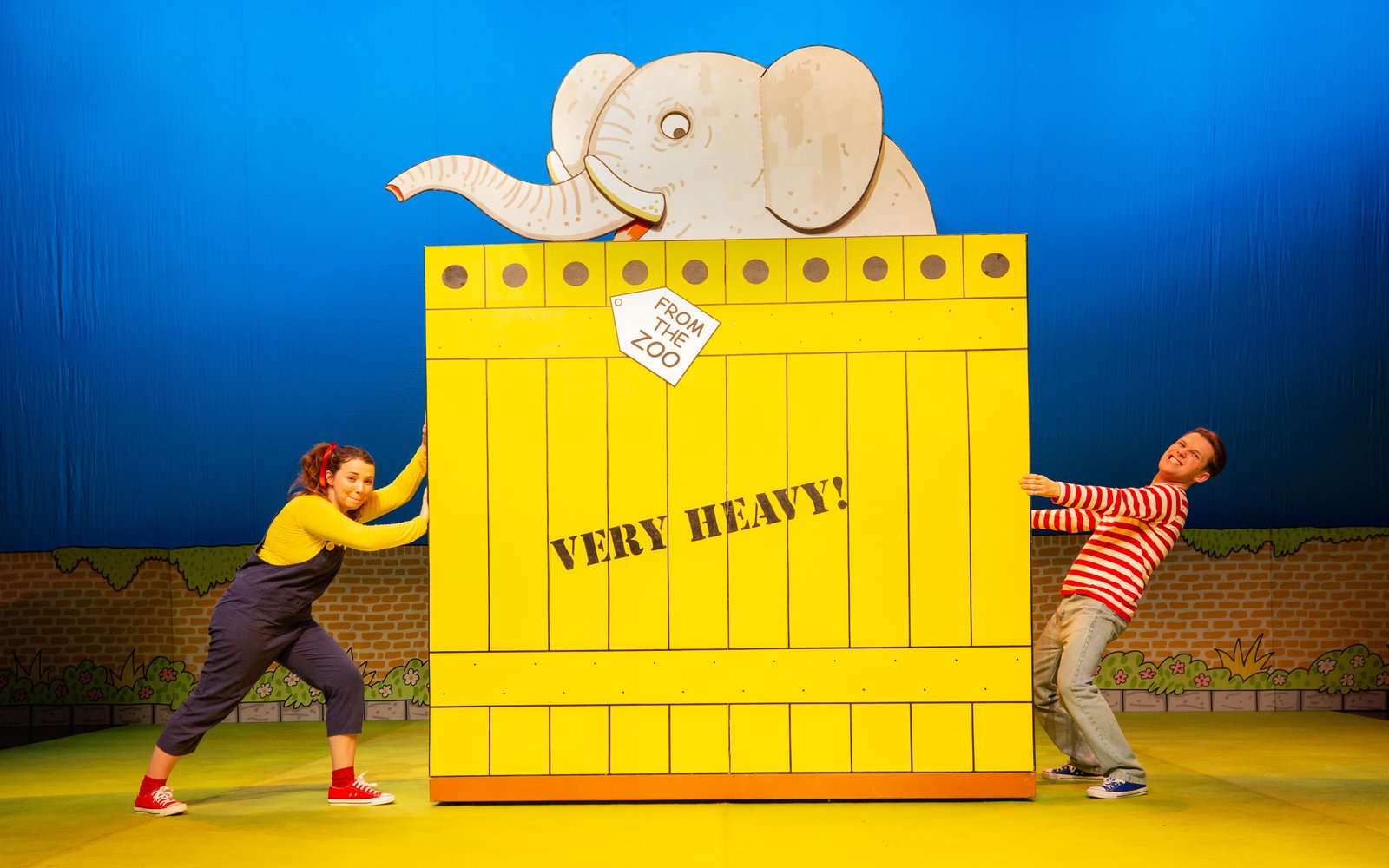A boy and a girl push a big yellow crate with an elephant inside it