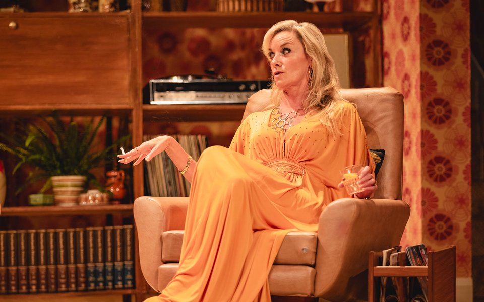 A blonde woman in a glamorous yellow dress sits in a living room arm chair