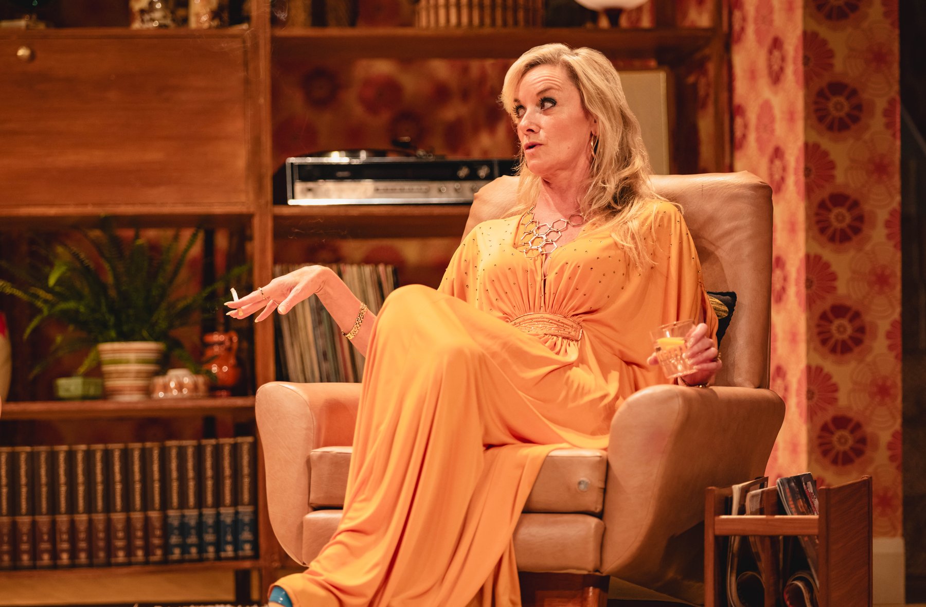 A blonde woman in a glamorous yellow dress sits in a living room arm chair
