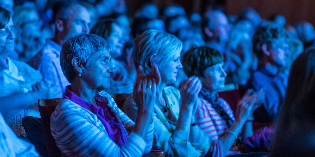 An audience bathed in blue light applaud.