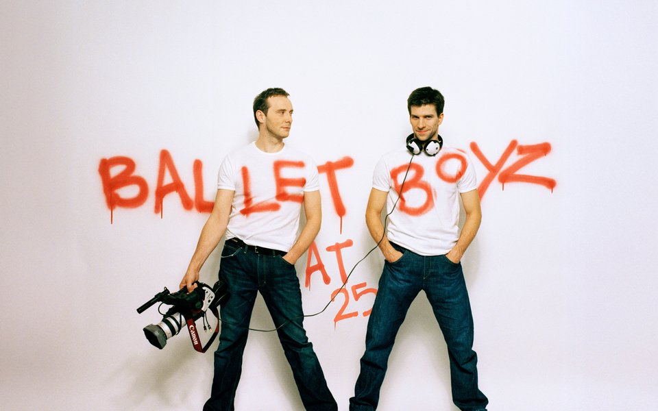 Two men wearing white tshirts and jeans stand against a white backdrop, they are carrying a video camera and there is graffiti writing across the wall spelling out 'BalletBoyz'