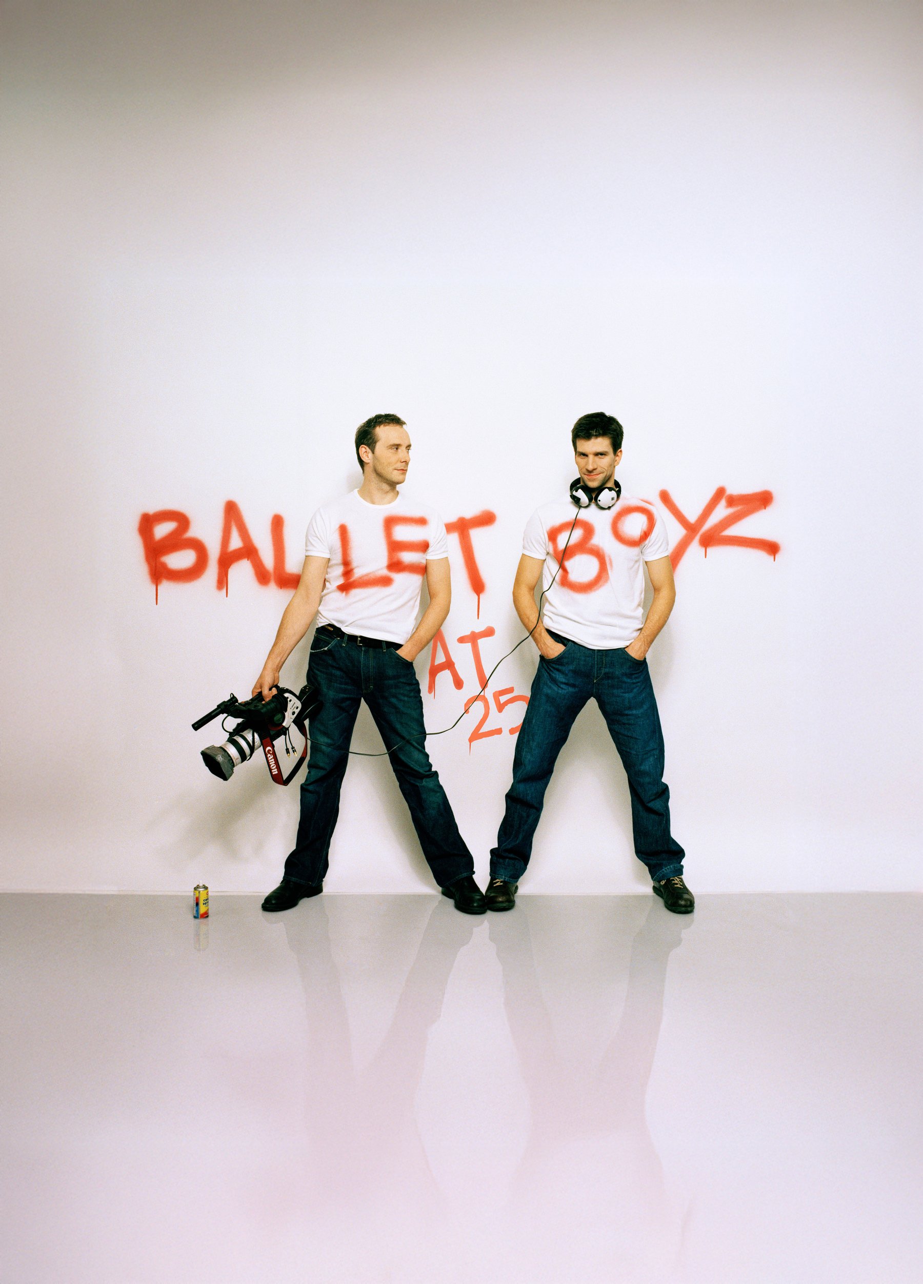 Two men wearing white tshirts and jeans stand against a white backdrop, they are carrying a video camera and there is graffiti writing across the wall spelling out 'BalletBoyz'