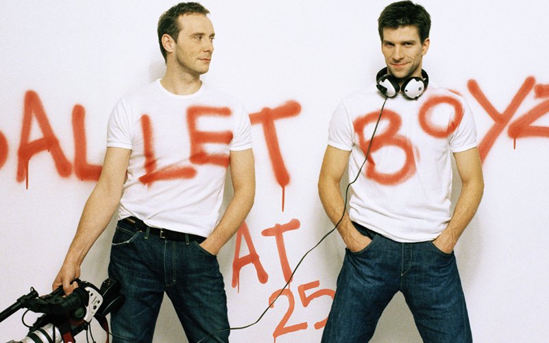 Two men wearing white tshirts and jeans stand against a white backdrop, they are carrying a video camera and there is graffiti writing across the wall spelling out 'BalletBoyz'