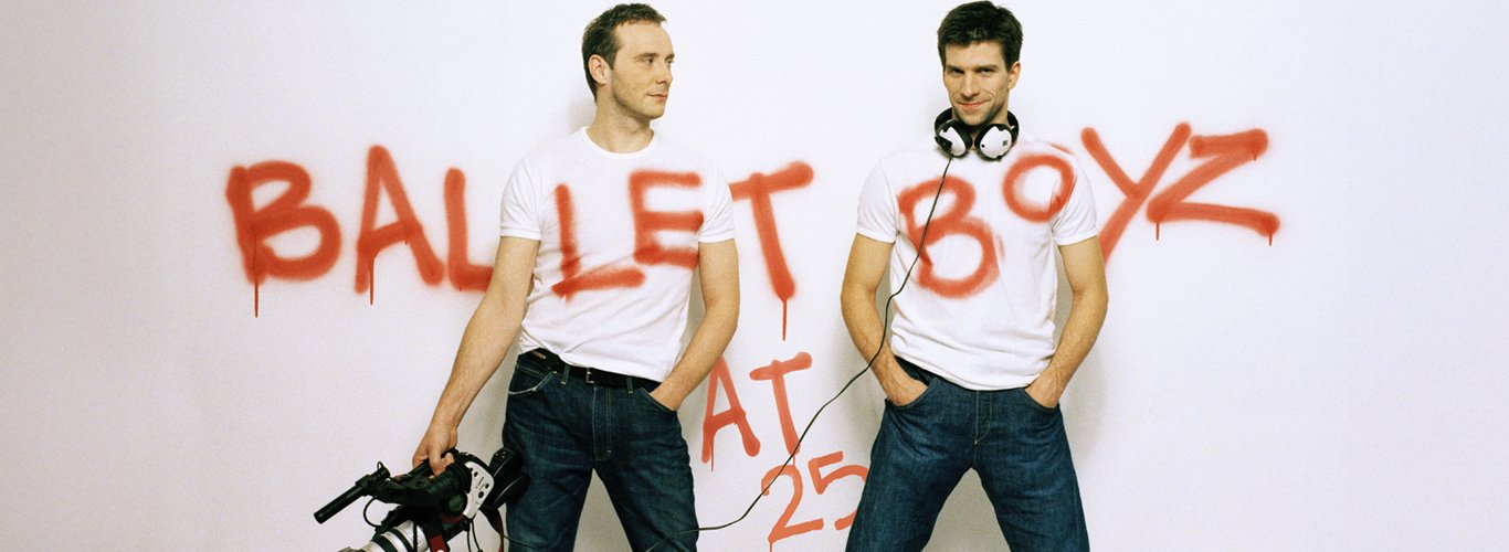 Two men wearing white tshirts and jeans stand against a white backdrop, they are carrying a video camera and there is graffiti writing across the wall spelling out 'BalletBoyz'