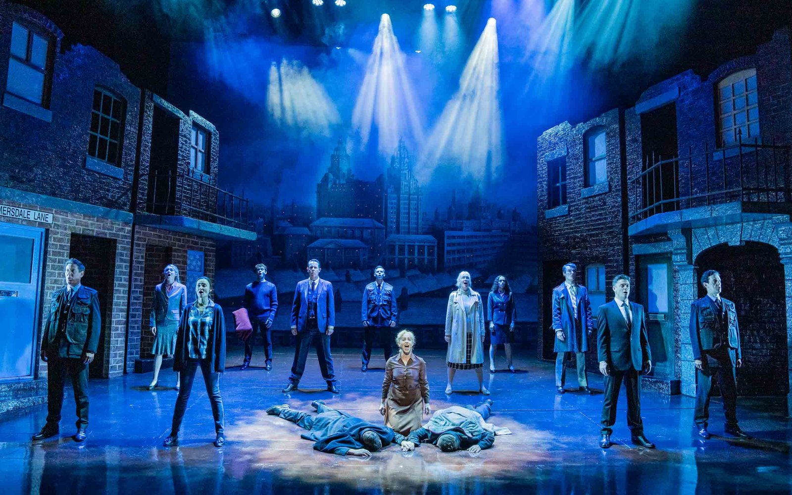 The cast stands spread out on the stage, facing outwards. In the middle, a woman kneels between two men who are lying face down.