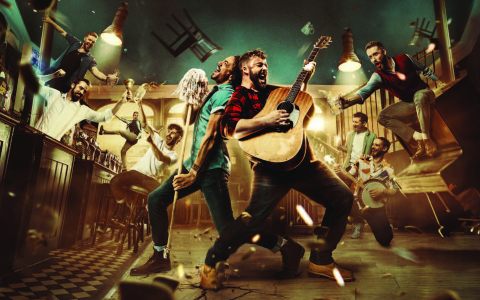 A chaotic scene inside a pub, chairs are flying, the tables and a piano is falling over. A man stands in the middle singing with a guitar.