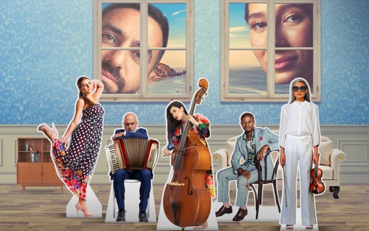 A group of paper cut out opera musicians stood together against a blue wall papered background, a man and a woman look through windows in the background