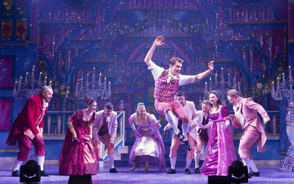 A group of people dance at a party, they are all wearing regal costumes in pink and purples and one man is leaping in the air as he dances