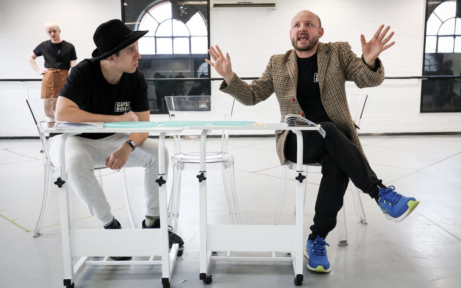 Two cast members sit at a table. One speaks and waves his hands while the other leans on the table and watches him.