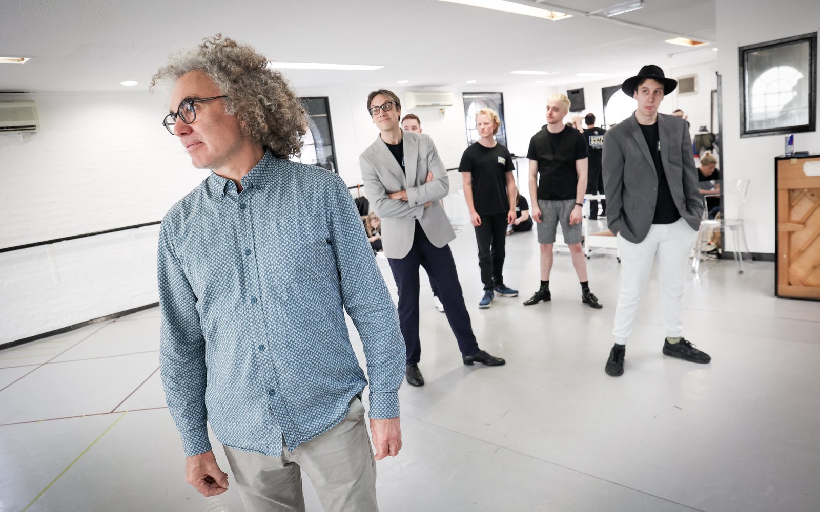 One actor in the foreground, looking to the side, while a group stand behind him, watching him.