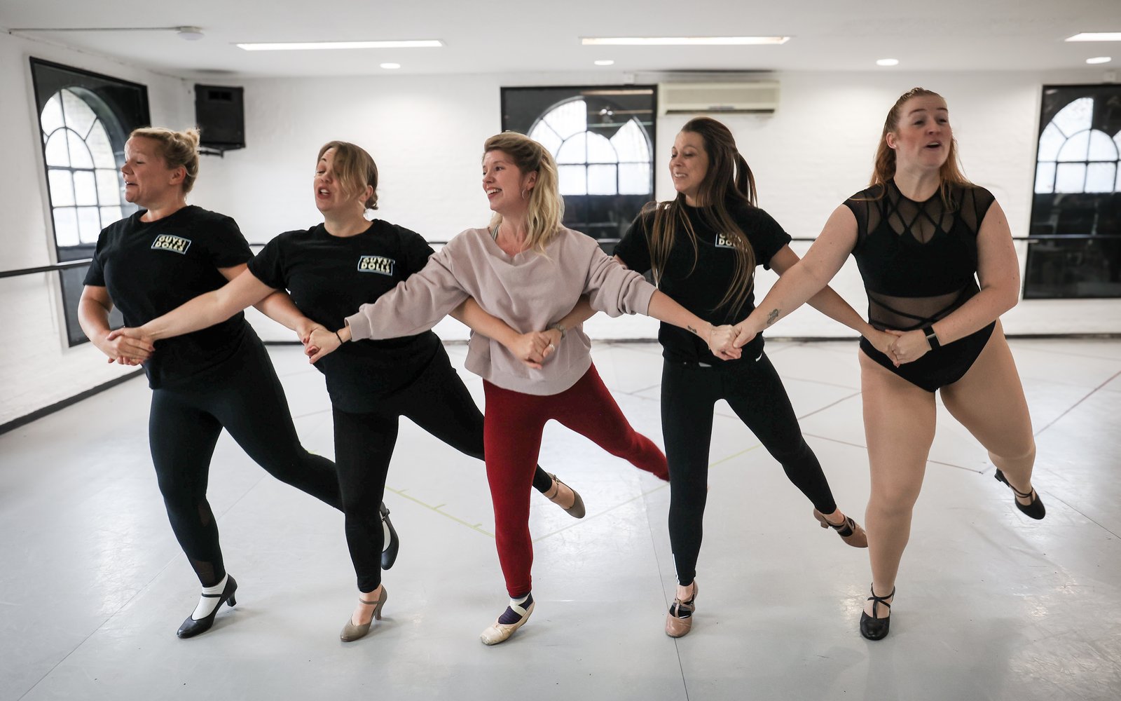 Five cast members mid-dance, interlocking arms in a line and kicking one foot out behind them.