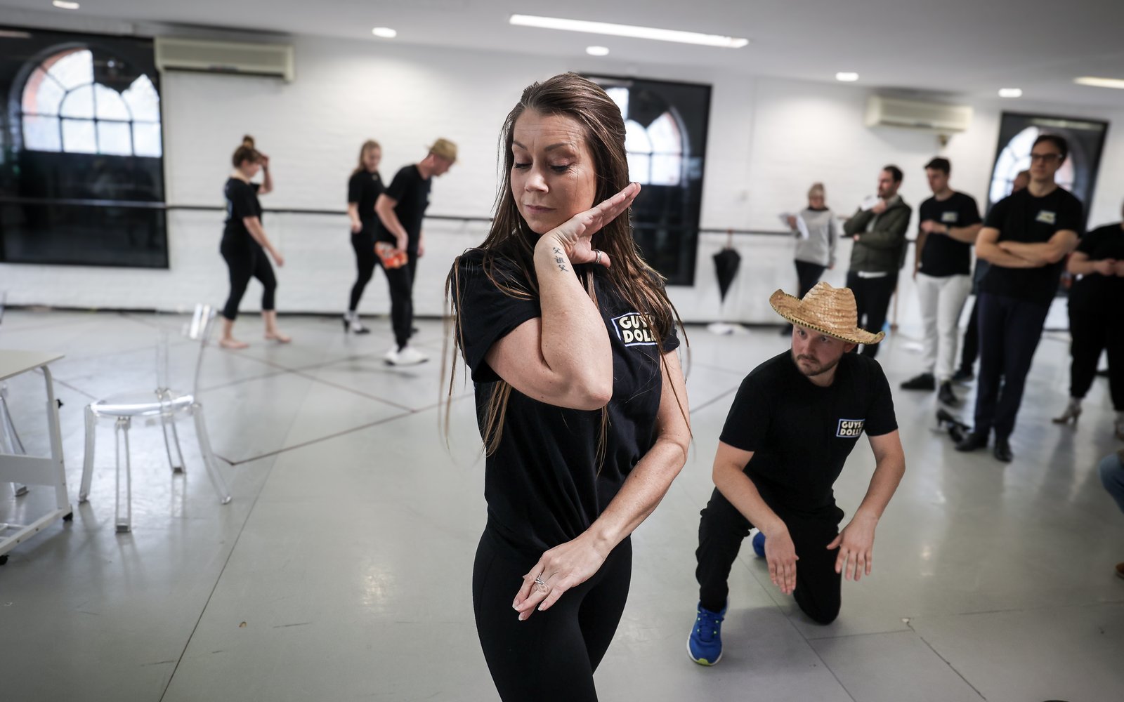An actor strikes a pose in the foreground, framing her face. Behind her, a man kneels on the floor watching her.