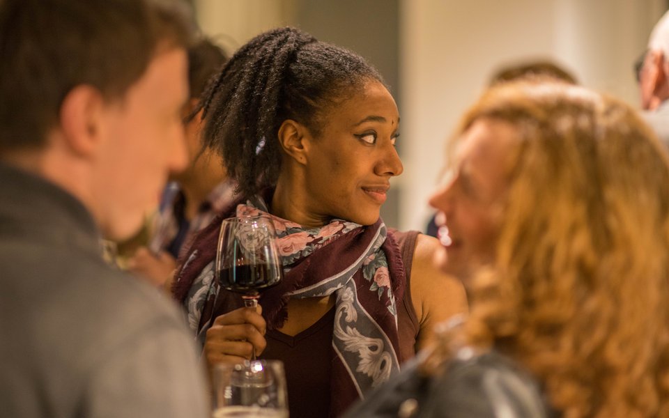 A lady stands in the centre of a busy room holding a glass of red wine.