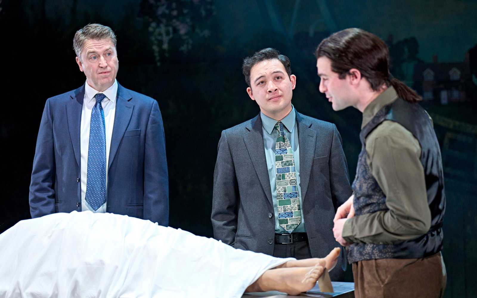 A body lies underneath a white sheet at a morgue, three men stand and look at it