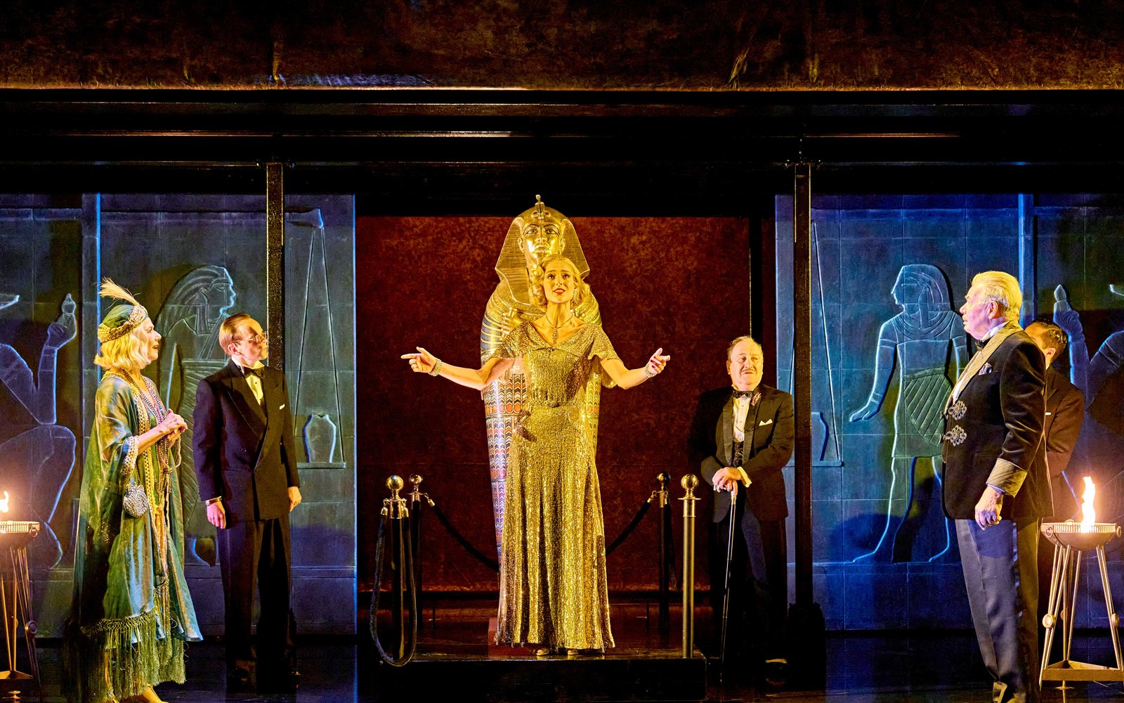 A woman in a gold sequin dress holds her arms out as she stands before an Egyptian coffin, a group of glamorous men and women watch her