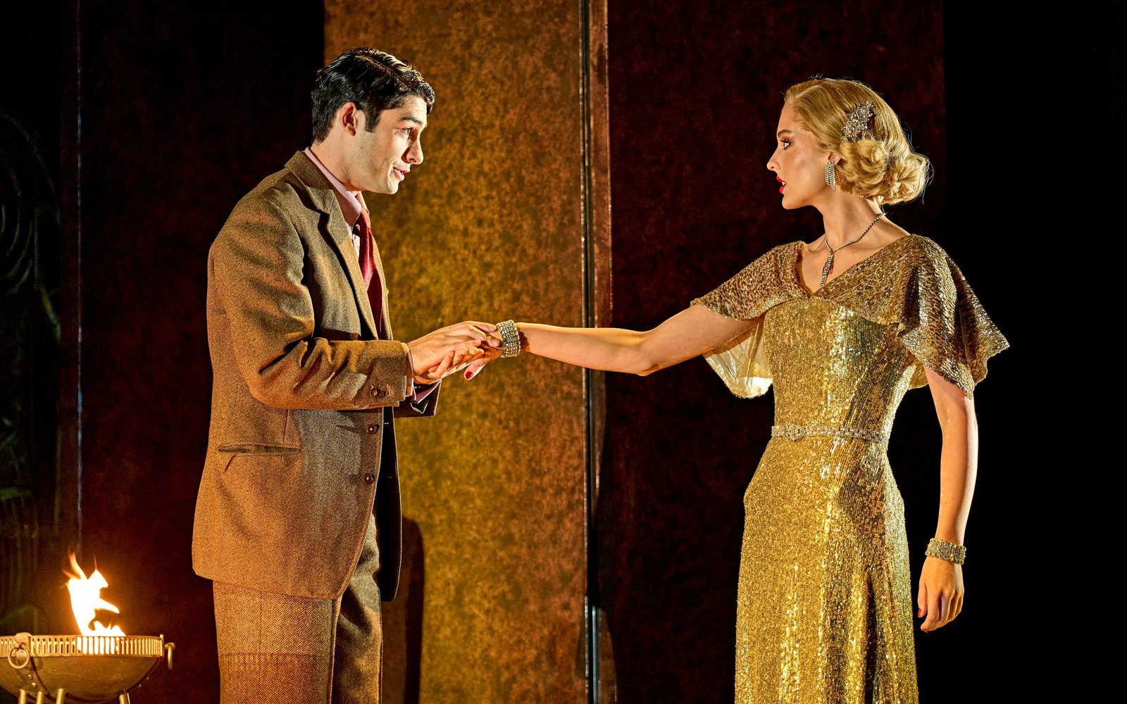 A man in a brown suit holds the outstretched hand of a blonde woman in a gold, 1920s-style gown.