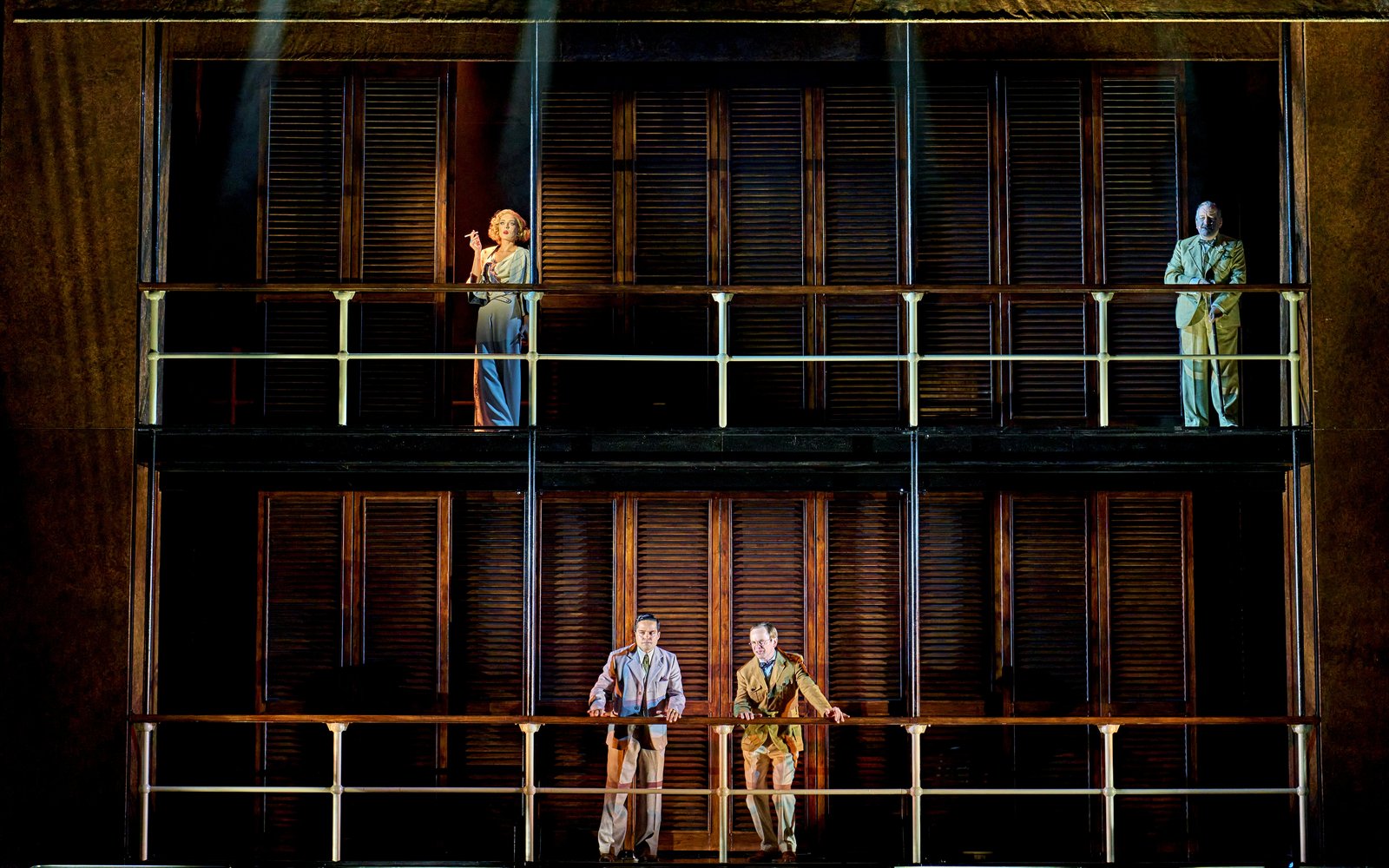 Four performers in 1920s-period clothing stand on two stacked balconies in front of dark wooden shutters, spaced apart.