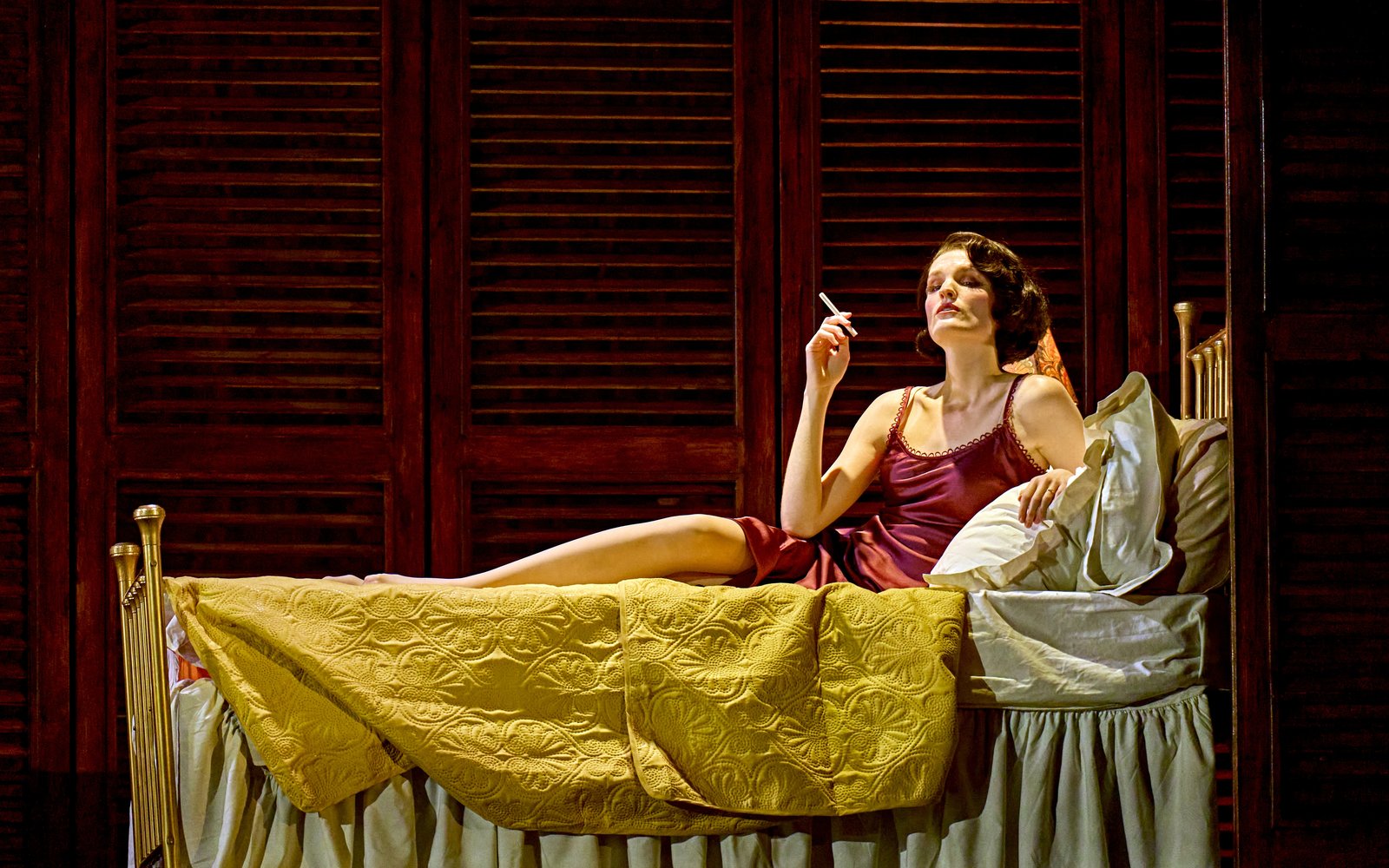 A woman in a vintage slip reclines on a bed, holding a cigarette and gazing upward.