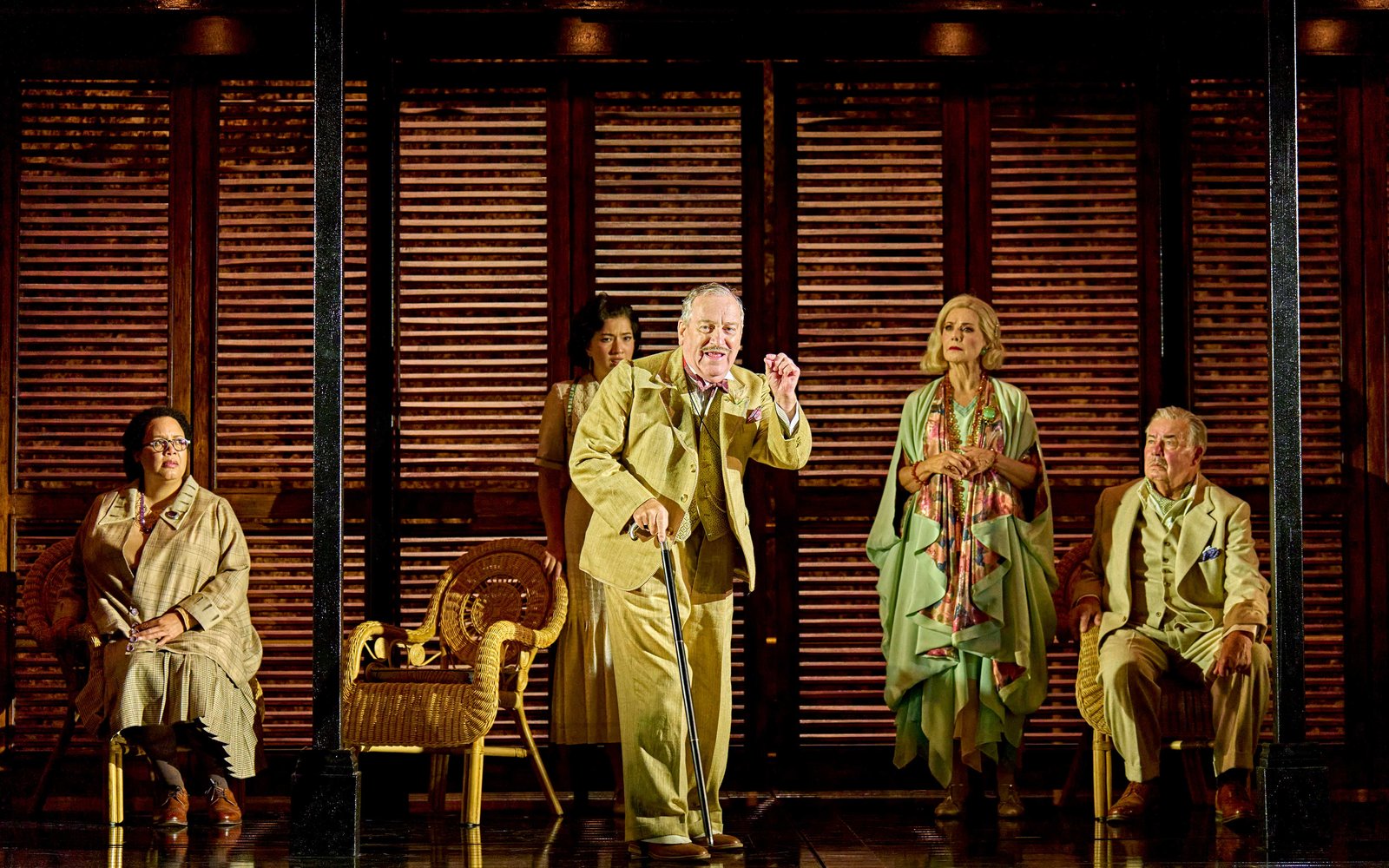 A man in a beige suit holding a cane talks animatedly, a group of people watch him intently