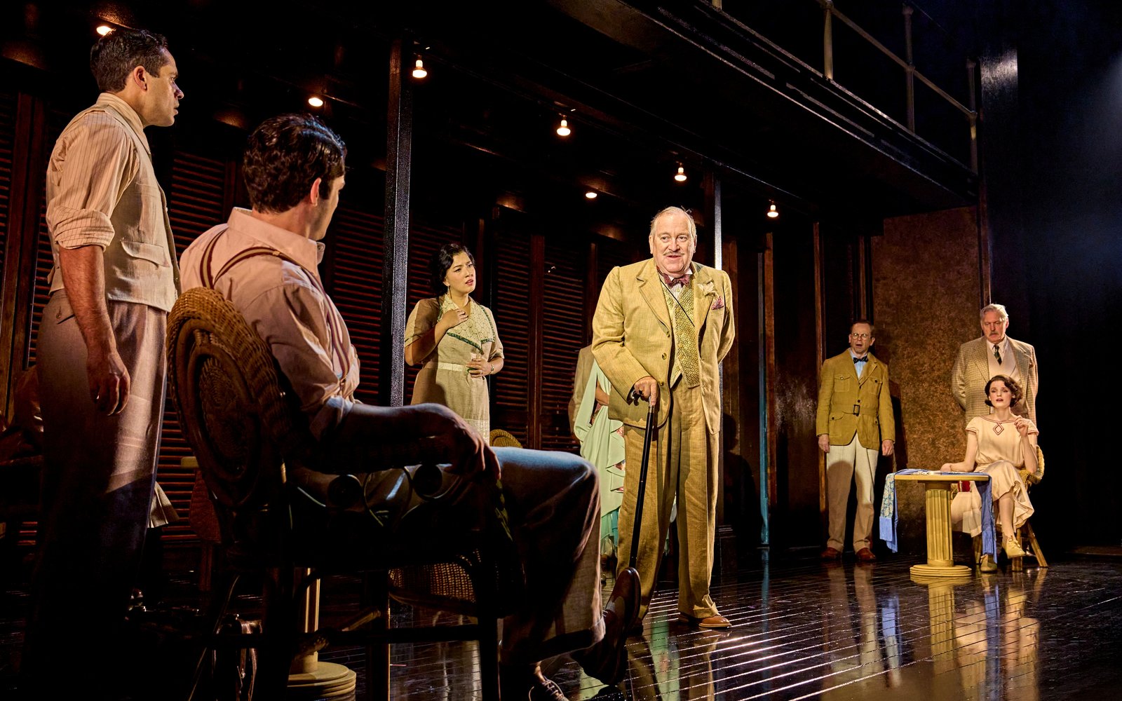 A man in a cream-coloured suit holding a cane addresses other characters surrounding him, all wearing 1920s-period clothing.