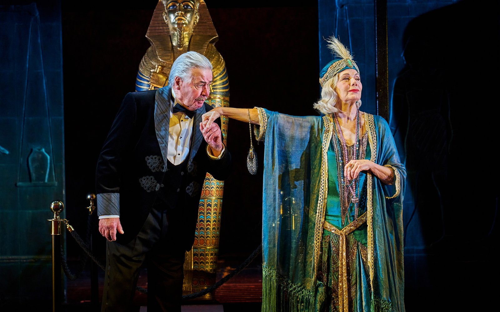 A man in a tuxedo bows to kiss the hand of an elegantly dressed woman in a flowing, Egyptian-inspired costume, with a golden pharaoh statue looming behind them.