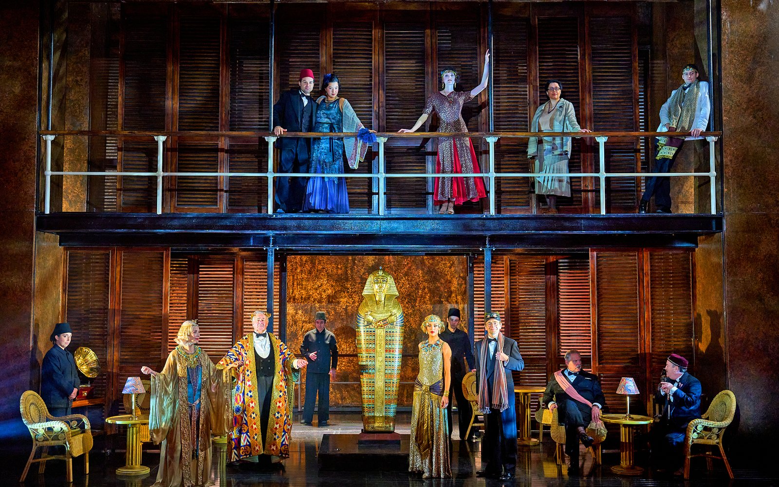 The whole cast, in period and Egyptian-inspired costumes. pose on a two-level set, with figures on a balcony above and a golden sarcophagus centered on the stage below.