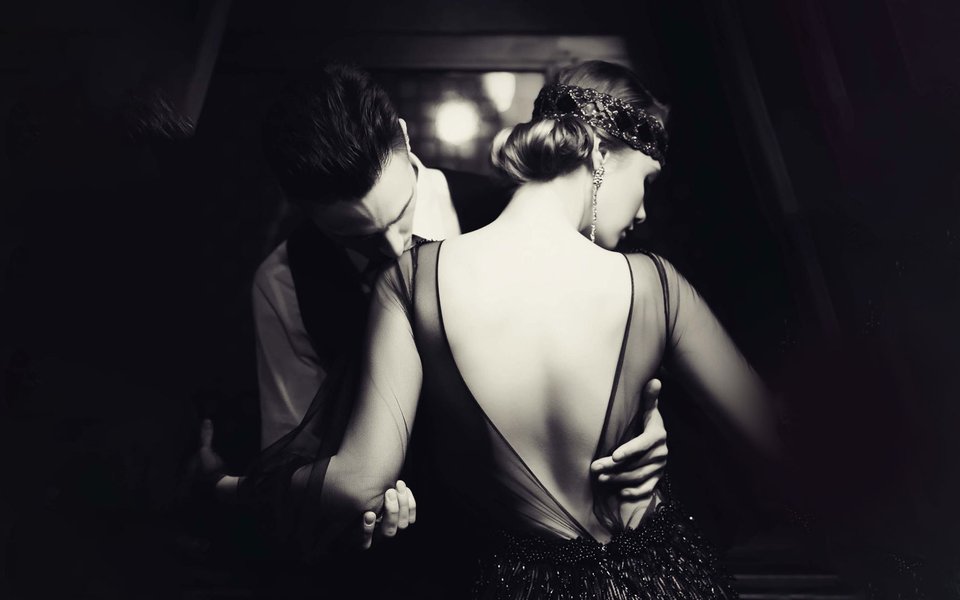 A black and white image of a glamorous 1920s man and a woman, the man is holding the woman and is kissing her shoulder as she looks away