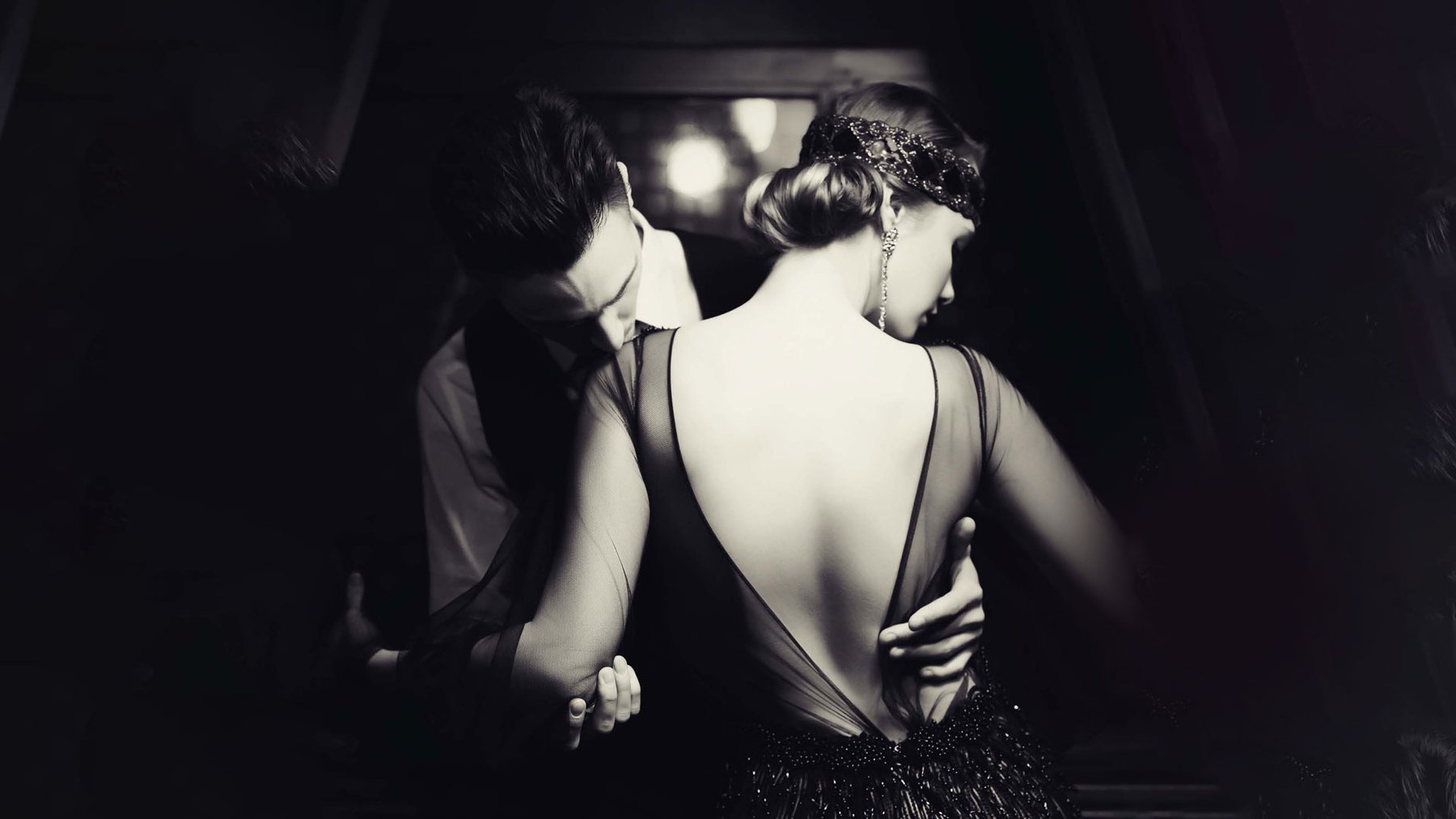 A black and white image of a glamorous 1920s man and a woman, the man is holding the woman and is kissing her shoulder as she looks away