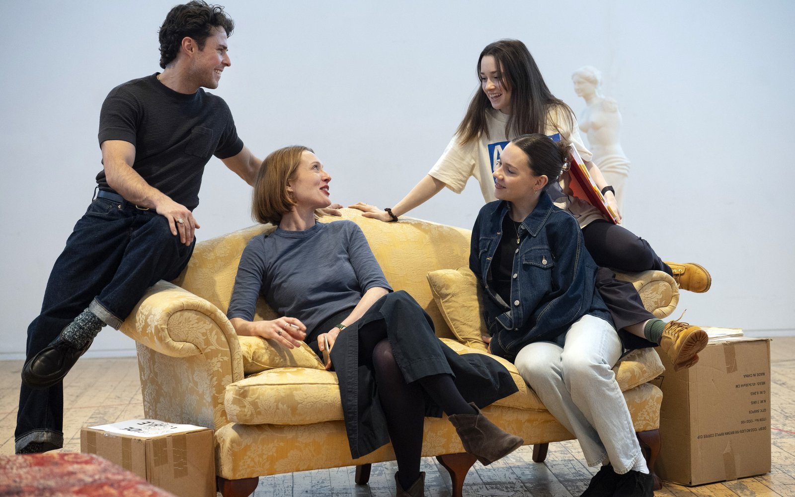 Three women and a man sit together on a yellow sofa, they are laughing and chatting happily