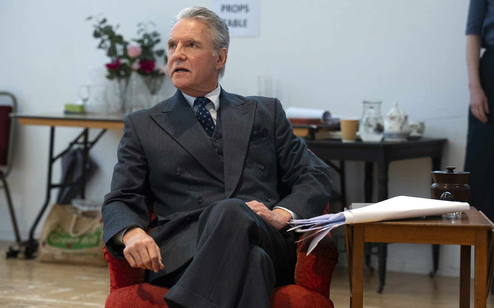 Michael Praed sits in a chair wearing a suit