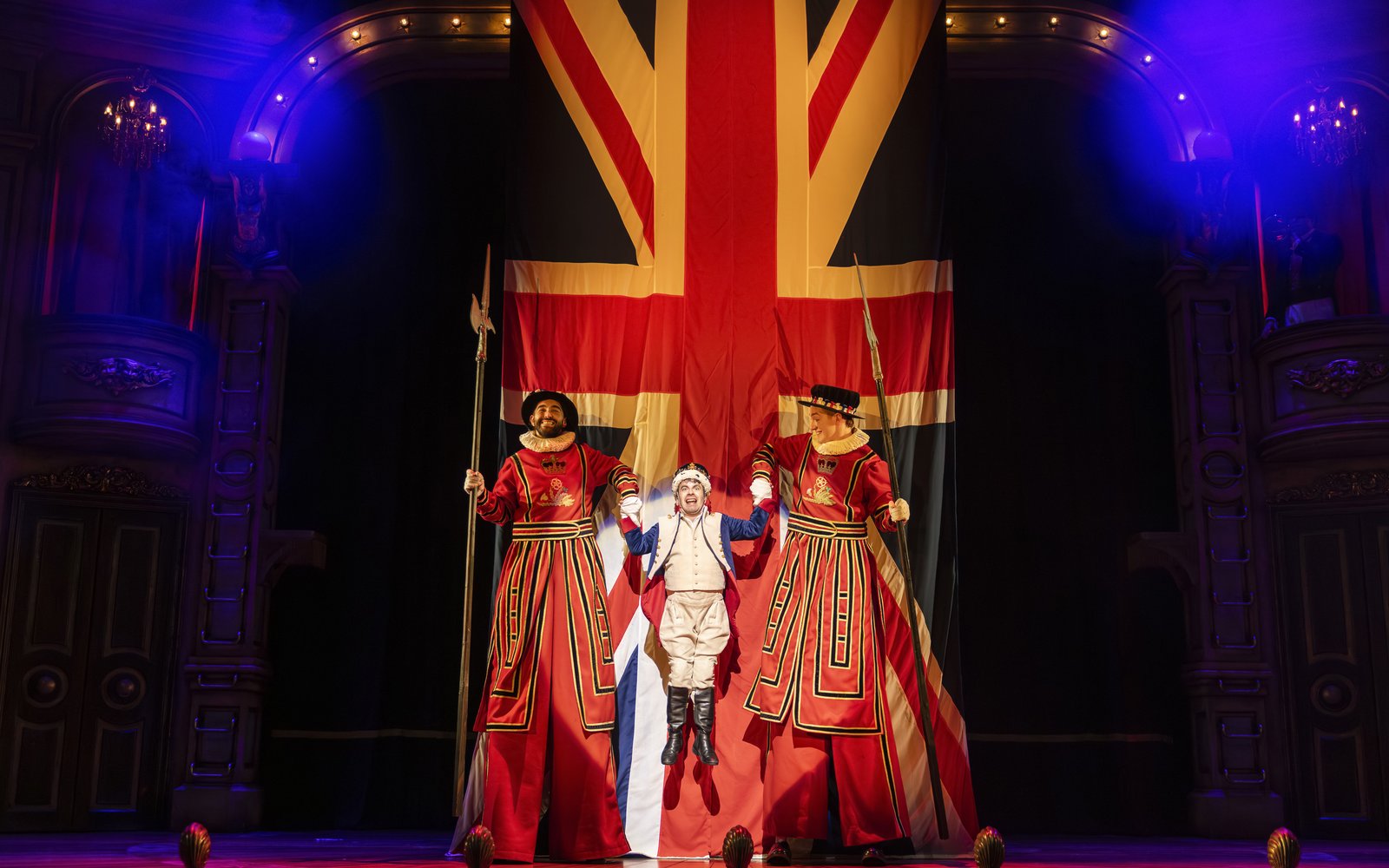 A man dressed as a British King is being pulled up by two people on stilts, there is a large Great British flag behind them
