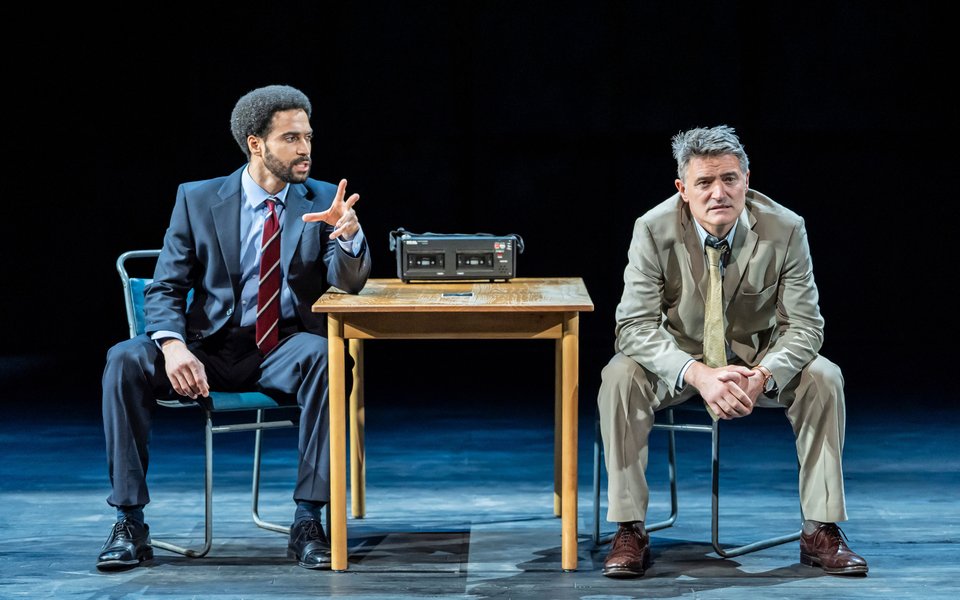 Two men sit either side of a wooden desk with a police recorder on, they are both wearing suits
