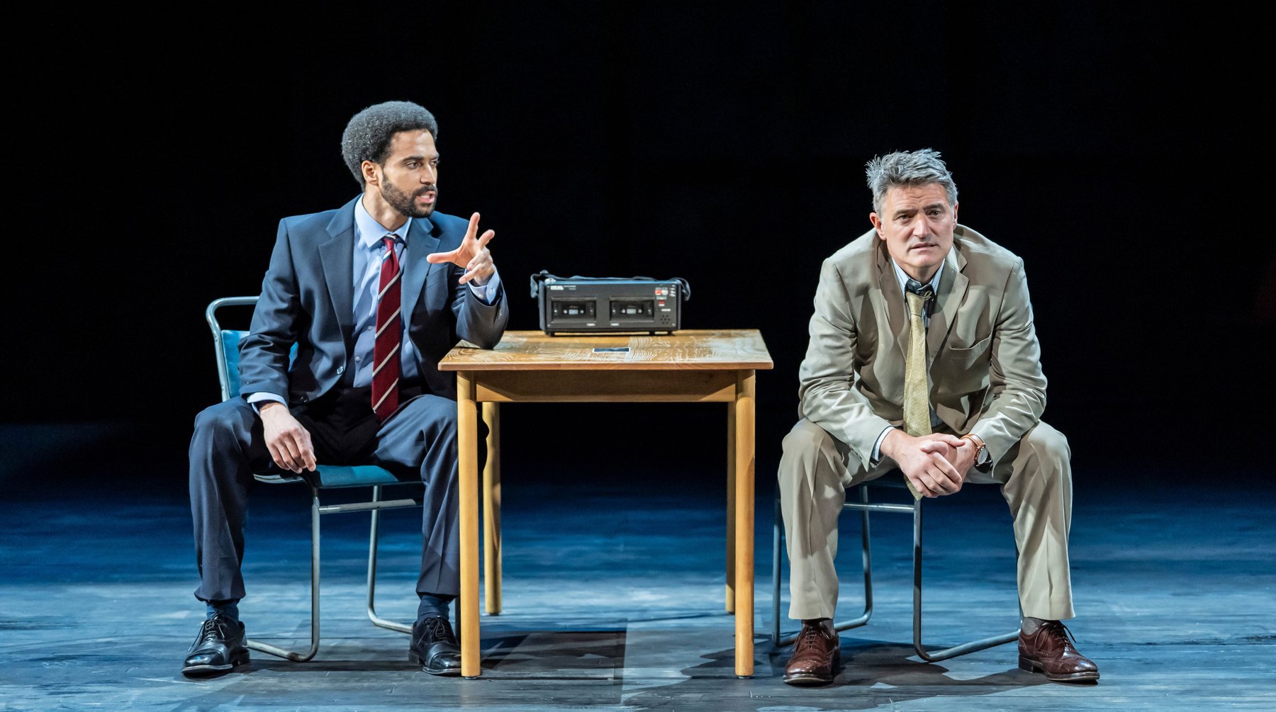 Two men sit either side of a wooden desk with a police recorder on, they are both wearing suits