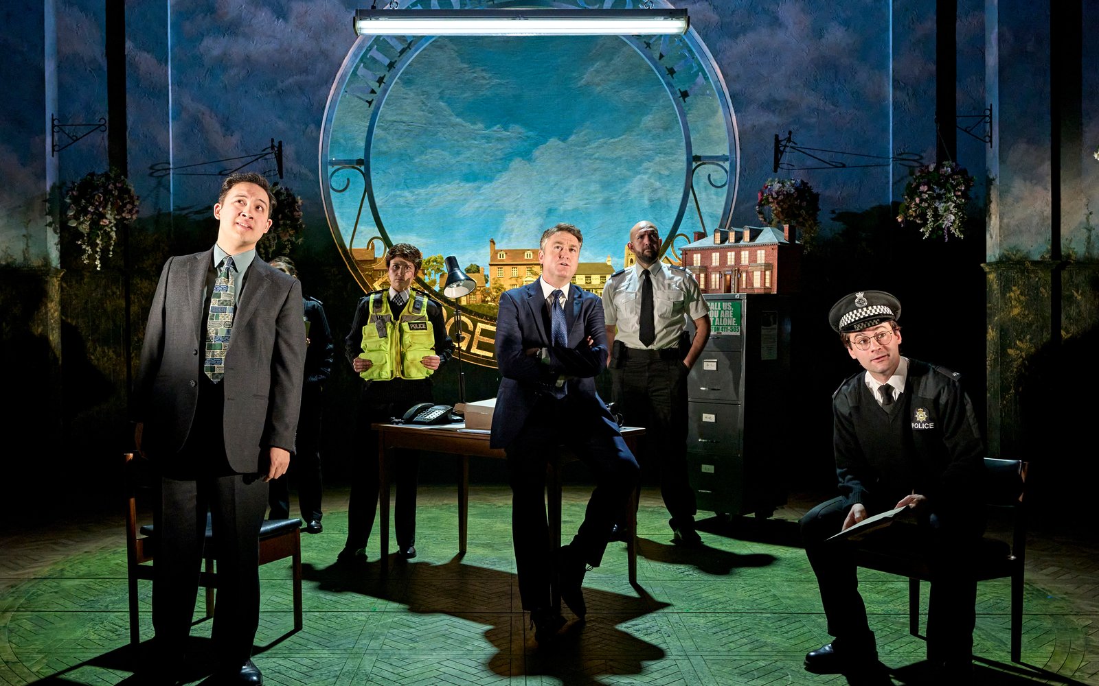 A group of men including policemen and men in suits stand together, there is the view of a picturesque village behind them