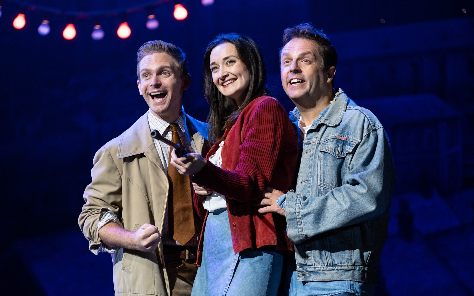 A woman holding a toy rifle stands between two men. They are all smiling.
