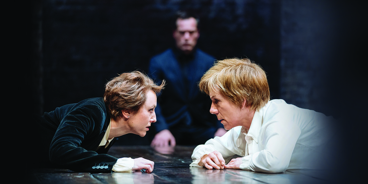 Two women lay on the floor on their front, they are staring into each other's eyes
